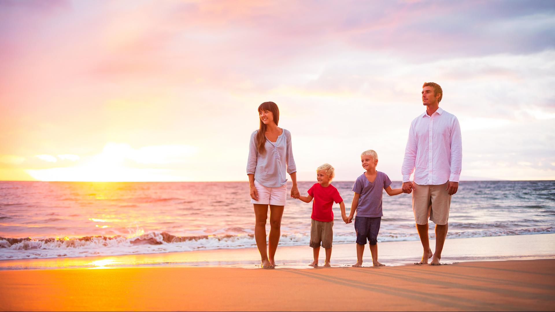 Famiglia cammina sulla spiaggia al tramonto, tenendosi per mano.
